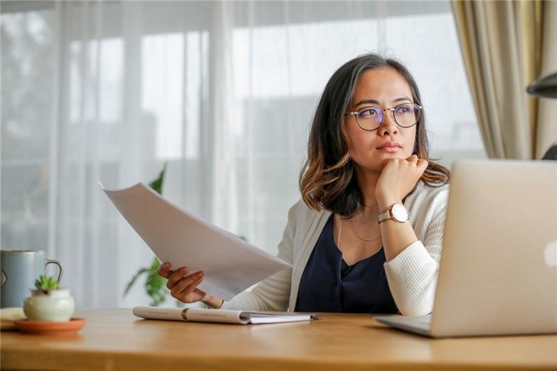 Seorang wanita merenungkan faktor penting saat memilih asuransi perjalanan Schengen sambil memegang dokumen di meja kerja modern dengan laptop dan tanaman hijau di latar belakang terang.