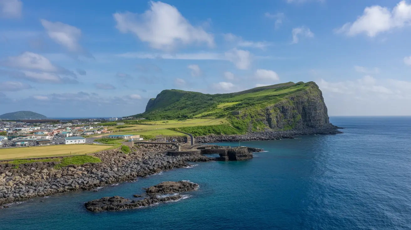 Pemandangan dramatis Tebing Seongsan Ilchulbong di Pulau Jeju yang hijau subur dengan rumput segar, bebatuan vulkanik hitam di tepi laut biru jernih, hamparan kampung kecil dan ladang di lereng, dikelilingi awan putih mengambang di langit cerah biru.